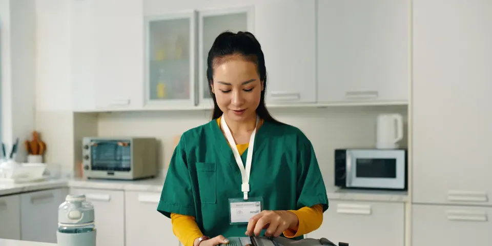 Healthcare professional packing a medical bag while standing in a contemporary kitchen setting with various kitchen appliances visible in background